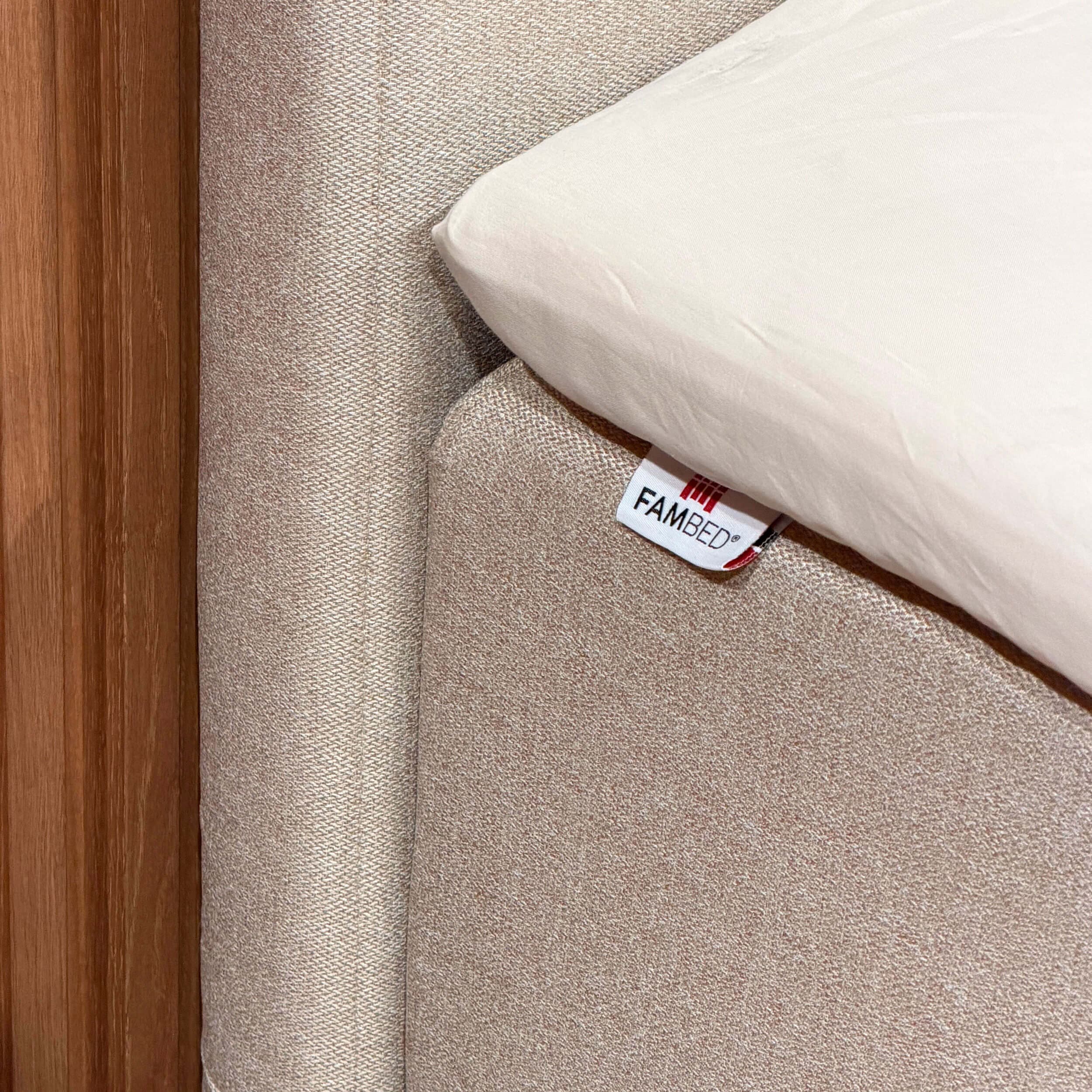 Close-up of the beige cover fitted around the corner of the bed, with a FAMBED label and white sheet on top